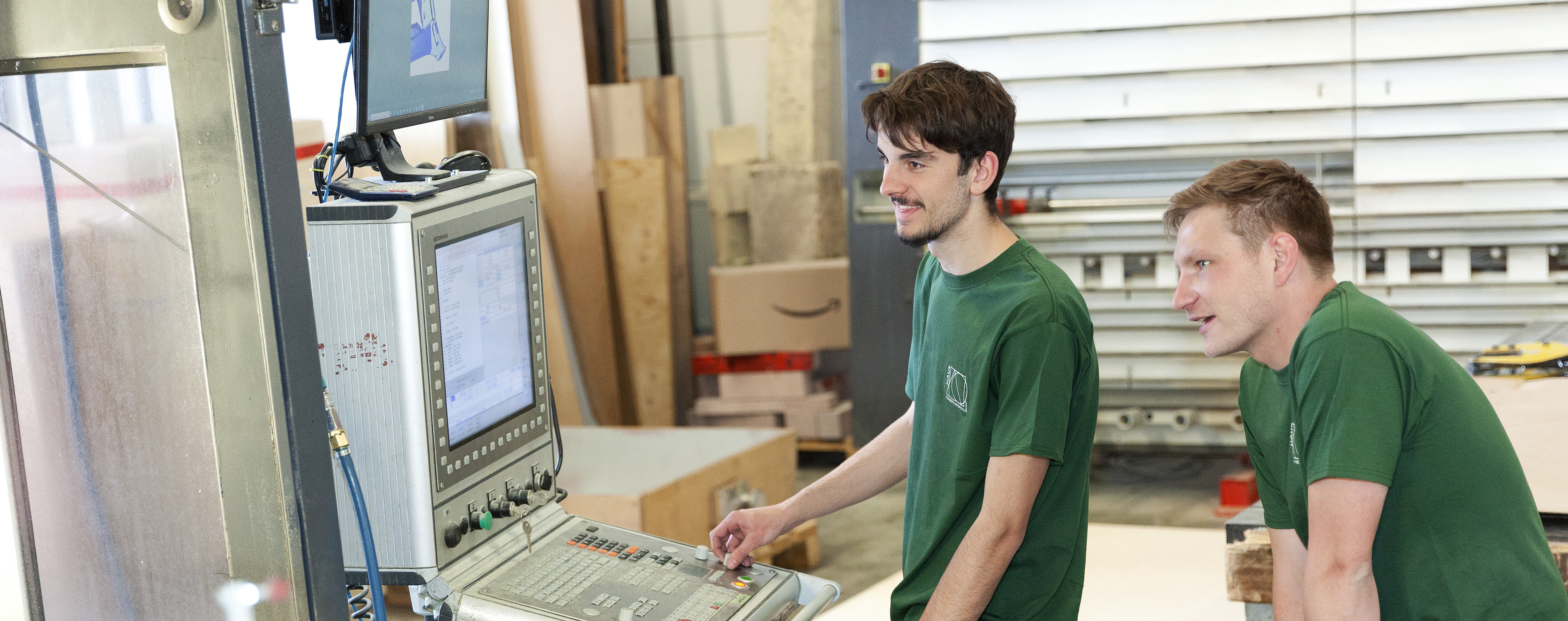 Zwei Männer in grünen T-Shirts stehen an einer Maschinensteuerung. Sie beobachten aufmerksam, während sie arbeiten. Holzmaterialien sind im Hintergrund sichtbar.