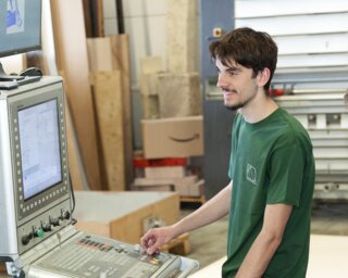 Zwei Männer in grünen T-Shirts stehen an einer Maschinensteuerung. Sie beobachten aufmerksam, während sie arbeiten. Holzmaterialien sind im Hintergrund sichtbar.