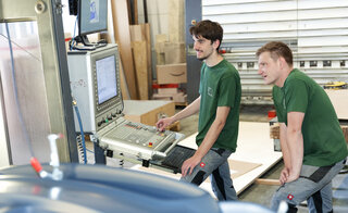 Zwei Männer in grünen T-Shirts stehen an einer Maschinensteuerung. Sie beobachten aufmerksam, während sie arbeiten. Holzmaterialien sind im Hintergrund sichtbar.