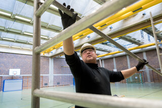 Ein Arbeiter in einer Halle baut ein Gerüst. Der Raum ist lichtdurchflutet.