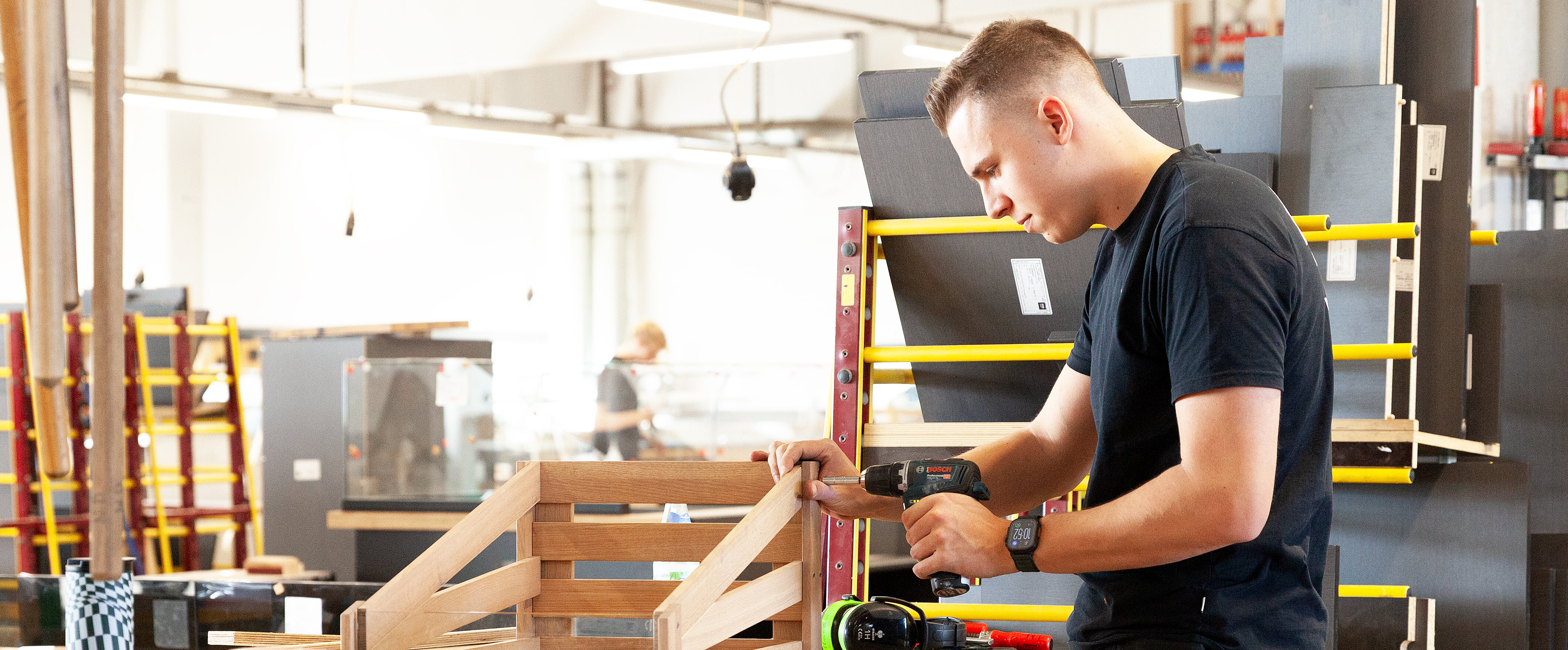 Ein junger Handwerker arbeitet in einer Werkstatt und konstruiert ein Holzprojekt mit einem Elektrowerkzeug. Im Hintergrund sind weitere Arbeitsstationen sichtbar.