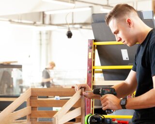 Ein junger Handwerker arbeitet in einer Werkstatt und konstruiert ein Holzprojekt mit einem Elektrowerkzeug. Im Hintergrund sind weitere Arbeitsstationen sichtbar.