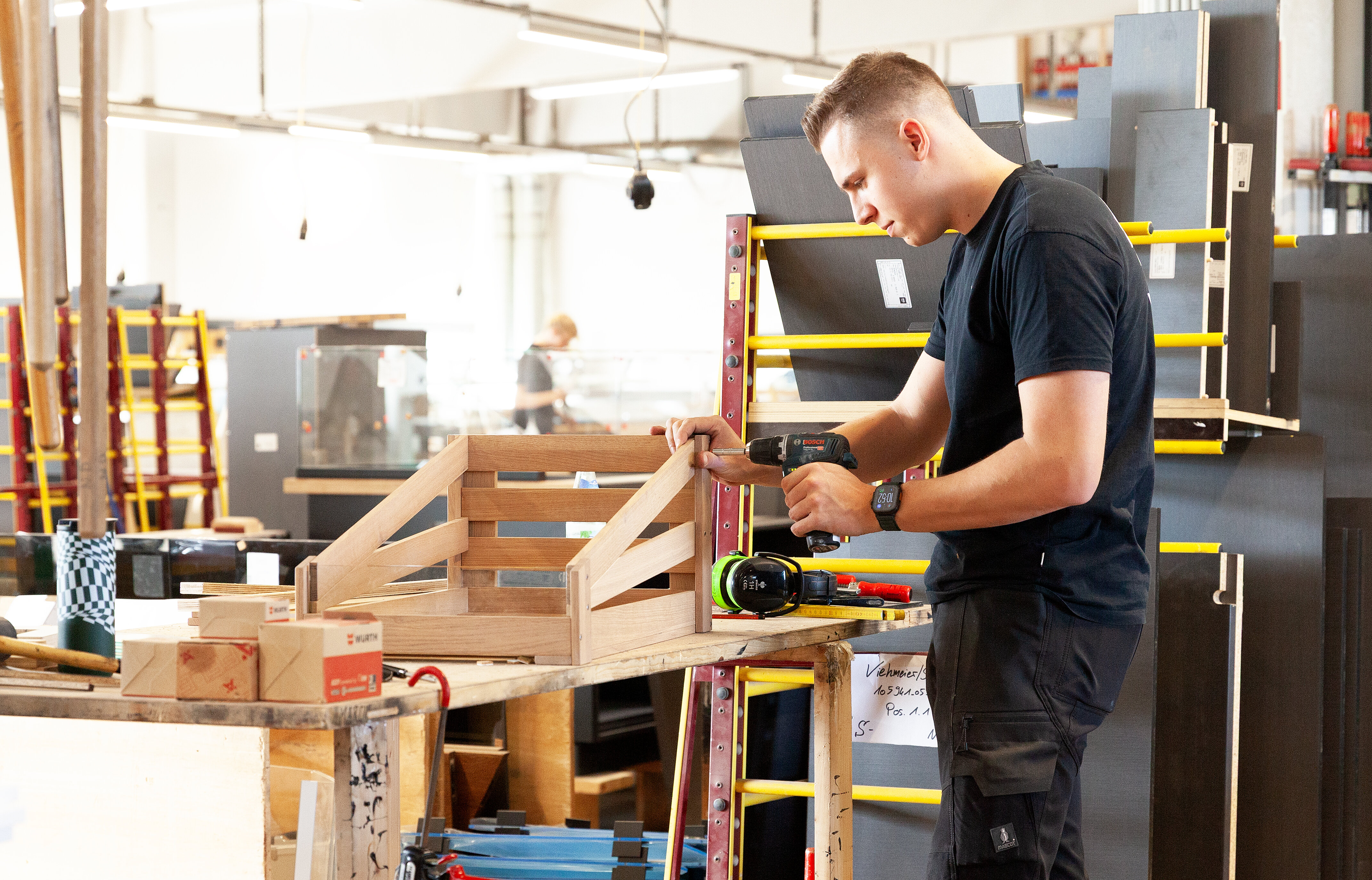 Ein junger Handwerker arbeitet in einer Werkstatt und konstruiert ein Holzprojekt mit einem Elektrowerkzeug. Im Hintergrund sind weitere Arbeitsstationen sichtbar.