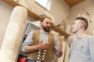 Zwei Männer stehen in einem Raum mit Skulpturen. Der erste spricht begeistert, während der andere aufmerksam zuhört.