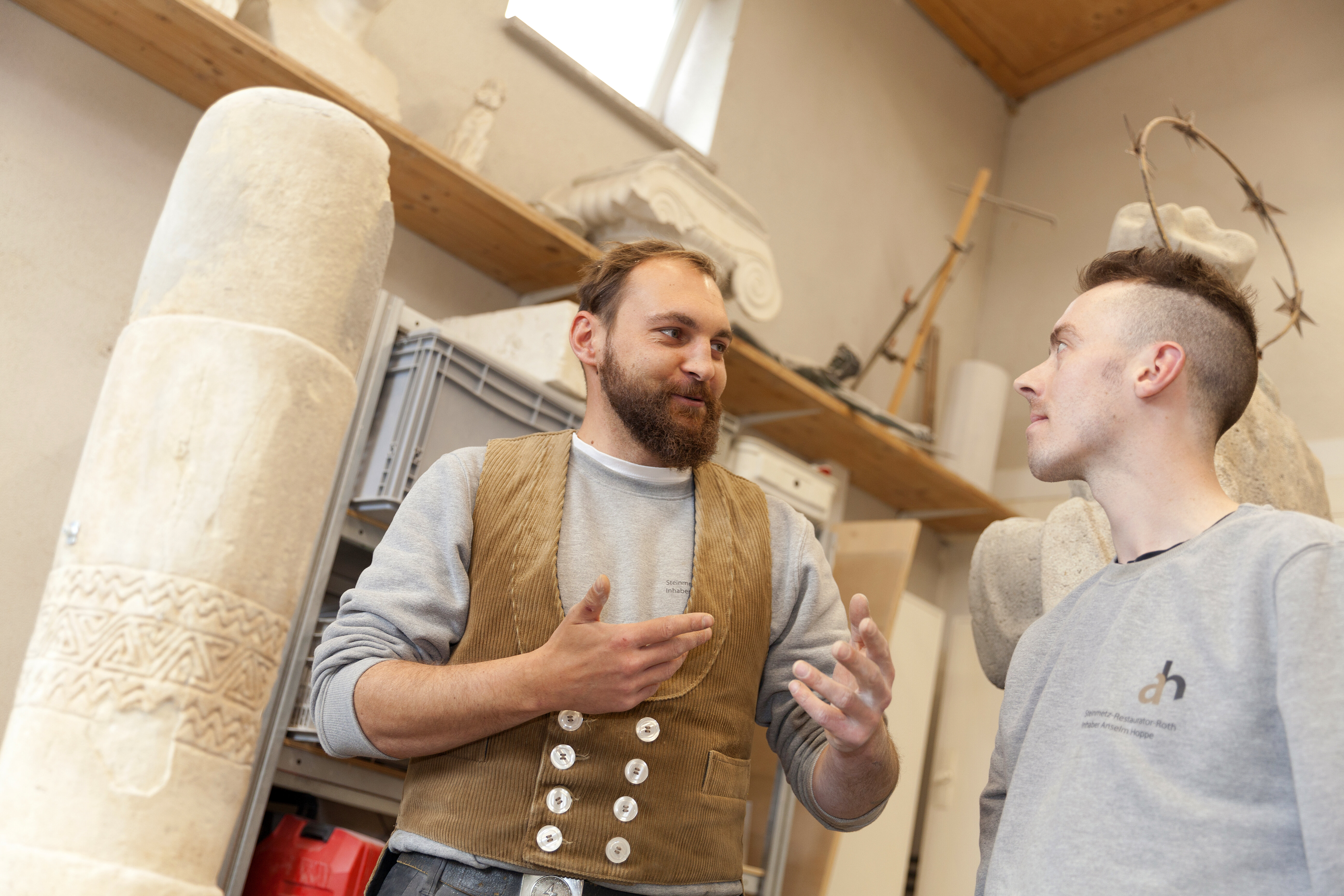 Zwei Männer stehen in einem Raum mit Skulpturen. Der erste spricht begeistert, während der andere aufmerksam zuhört.