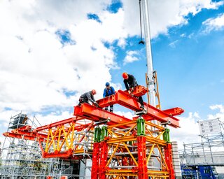 Auf der Baustelle arbeiten mehrere Personen an einem farbenfrohen Gerüst. Der Himmel ist blau mit vereinzelten Wolken.