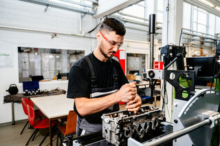 Ein junger Mann trägt Schutzbrille und arbeitet konzentriert an einem Motorblock in einer modernen Werkstatt. Werkbänke und Ausrüstung im Hintergrund.