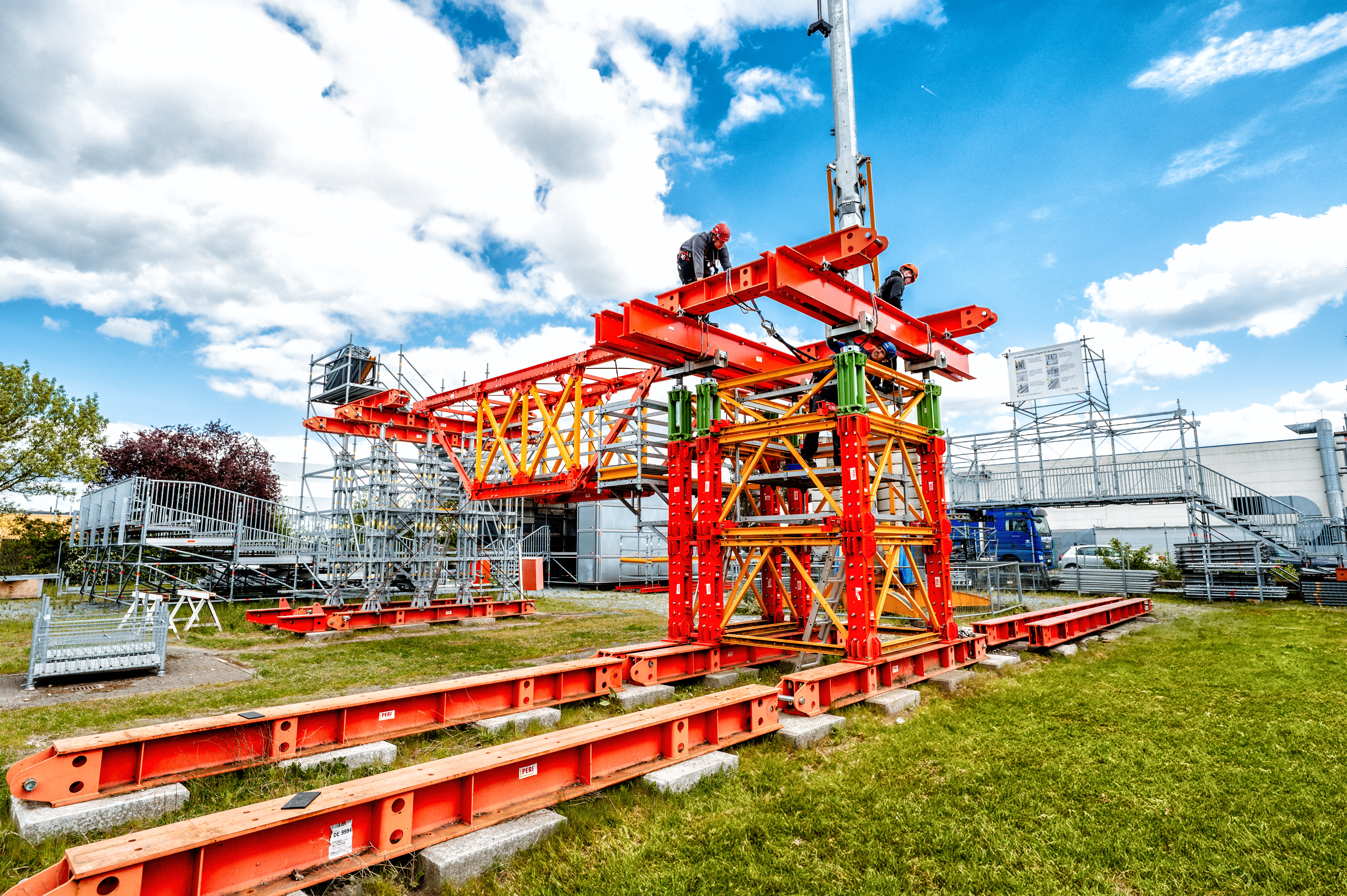 Das Bild zeigt ein orangefarbenes Baugerüst auf einer Wiese, mit Handwerkern, die an der Konstruktion arbeiten, und einem blauen Himmel im Hintergrund.