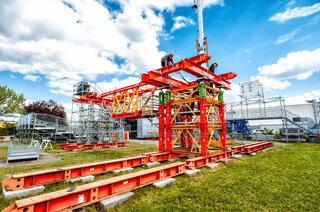 Das Bild zeigt ein orangefarbenes Baugerüst auf einer Wiese, mit Handwerkern, die an der Konstruktion arbeiten, und einem blauen Himmel im Hintergrund.