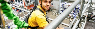 Ein junger Mann in einem Gelb trägt einen Helm und Sicherheitsgurt, während er auf einem Gerüst arbeitet. Die Umgebung ist industriell und gut ausgestattet.