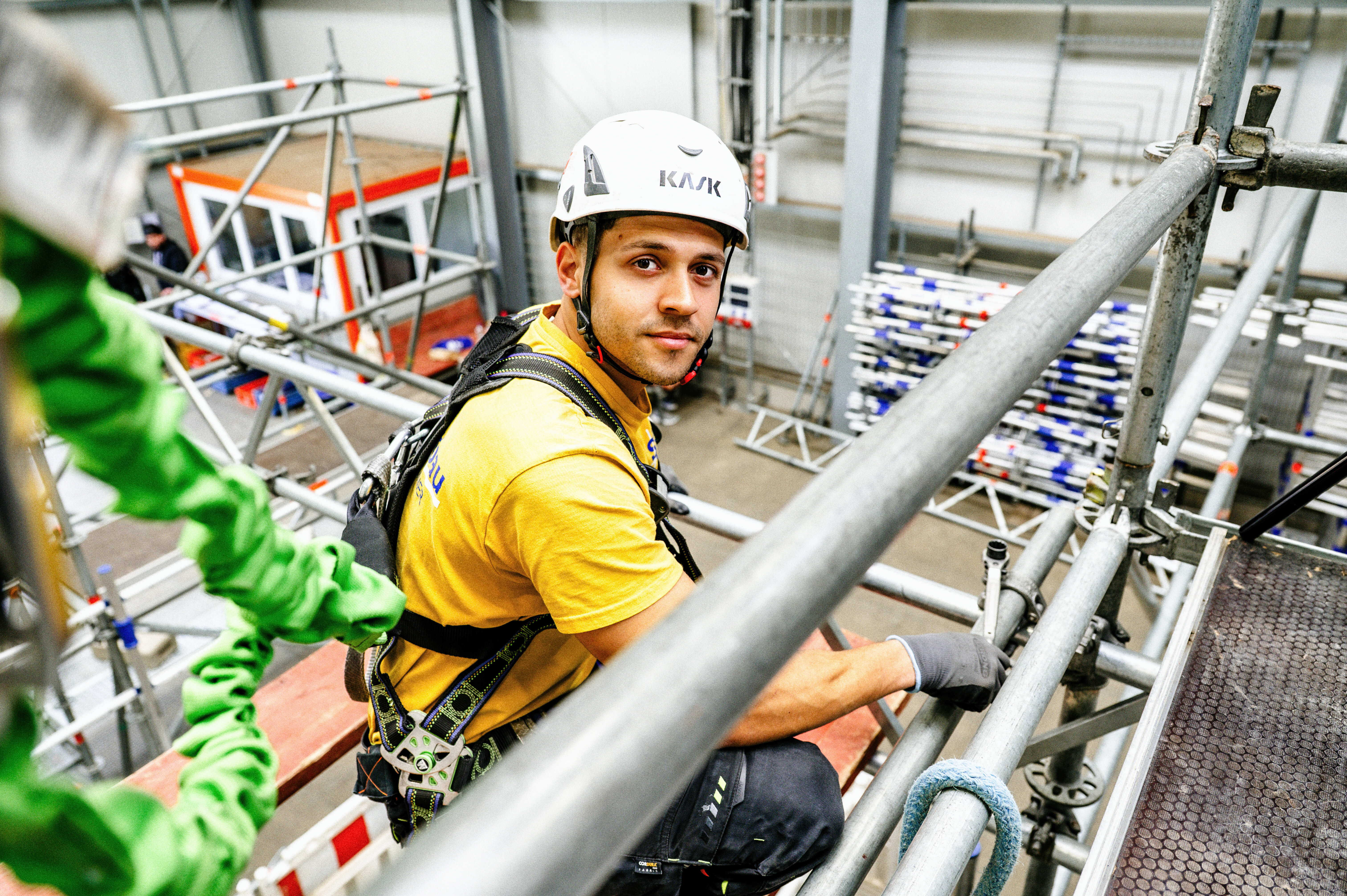 Ein junger Mann in einem Gelb trägt einen Helm und Sicherheitsgurt, während er auf einem Gerüst arbeitet. Die Umgebung ist industriell und gut ausgestattet.