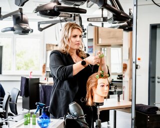 Eine Friseurin arbeitet konzentriert an einem Modellkopf mit Lockenwicklern in einem modernen Salon. Die Umgebung wirkt professionell und gut ausgestattet.