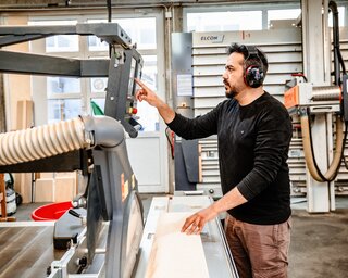 Der Handwerker arbeitet in einer Holzwerkstatt. Er trägt Gehörschutz und konzentriert sich auf eine Holzmaschine, während er das Material positioniert.