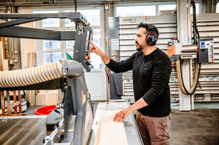 Der Handwerker arbeitet in einer Holzwerkstatt. Er trägt Gehörschutz und konzentriert sich auf eine Holzmaschine, während er das Material positioniert.