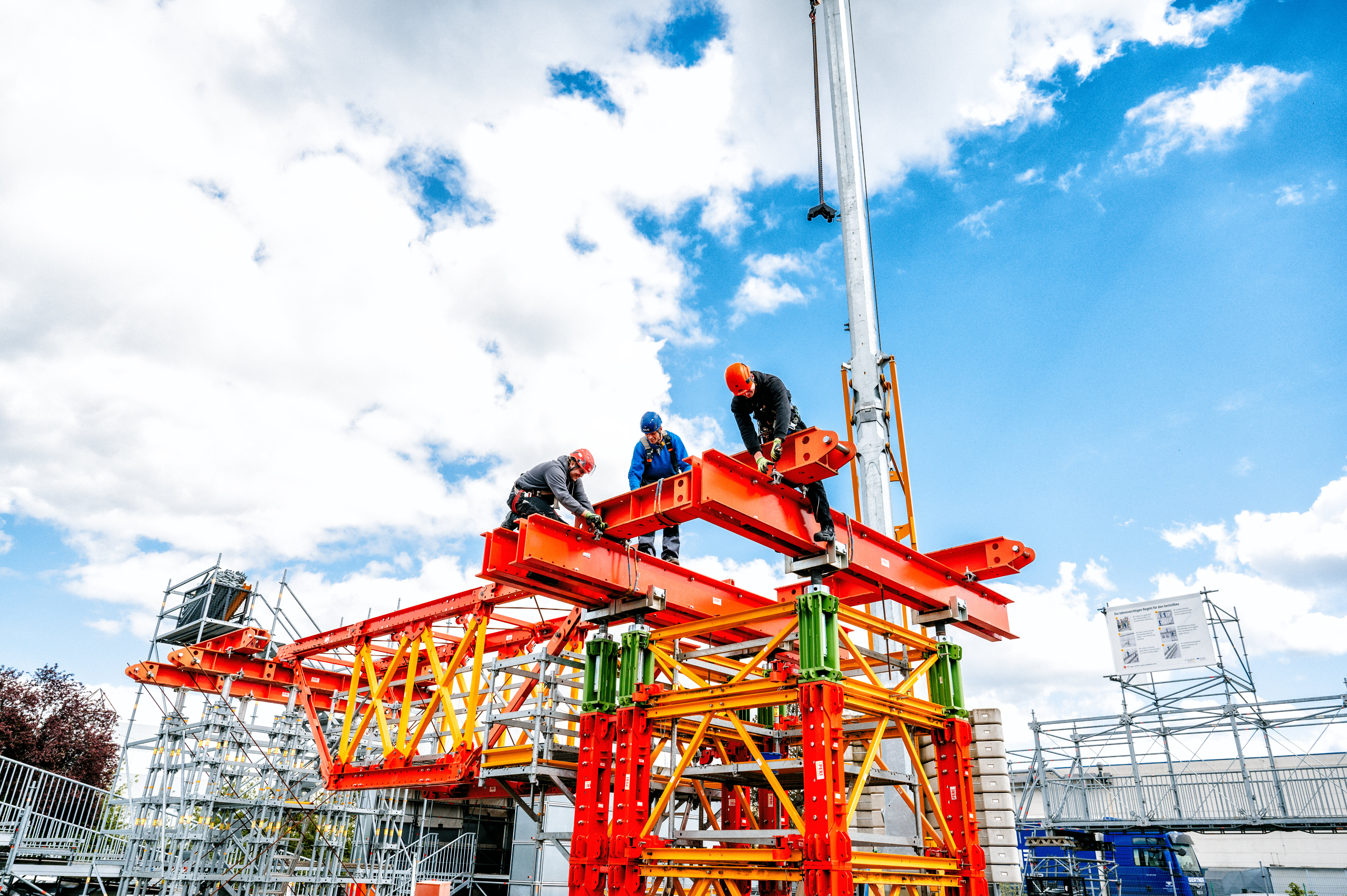 Auf der Baustelle arbeiten mehrere Personen an einem farbenfrohen Gerüst. Der Himmel ist blau mit vereinzelten Wolken.