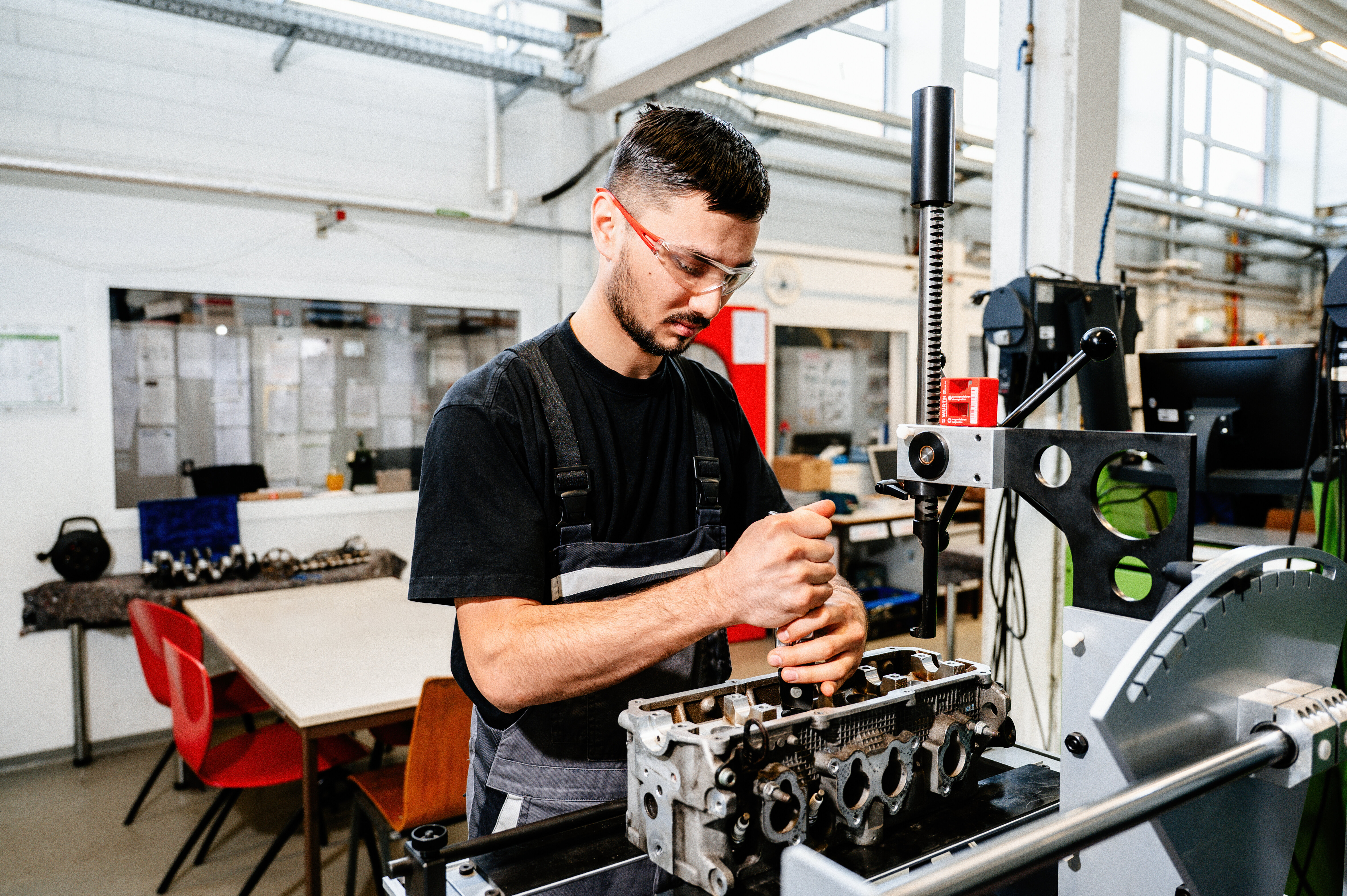 Ein junger Mann trägt Schutzbrille und arbeitet konzentriert an einem Motorblock in einer modernen Werkstatt. Werkbänke und Ausrüstung im Hintergrund.