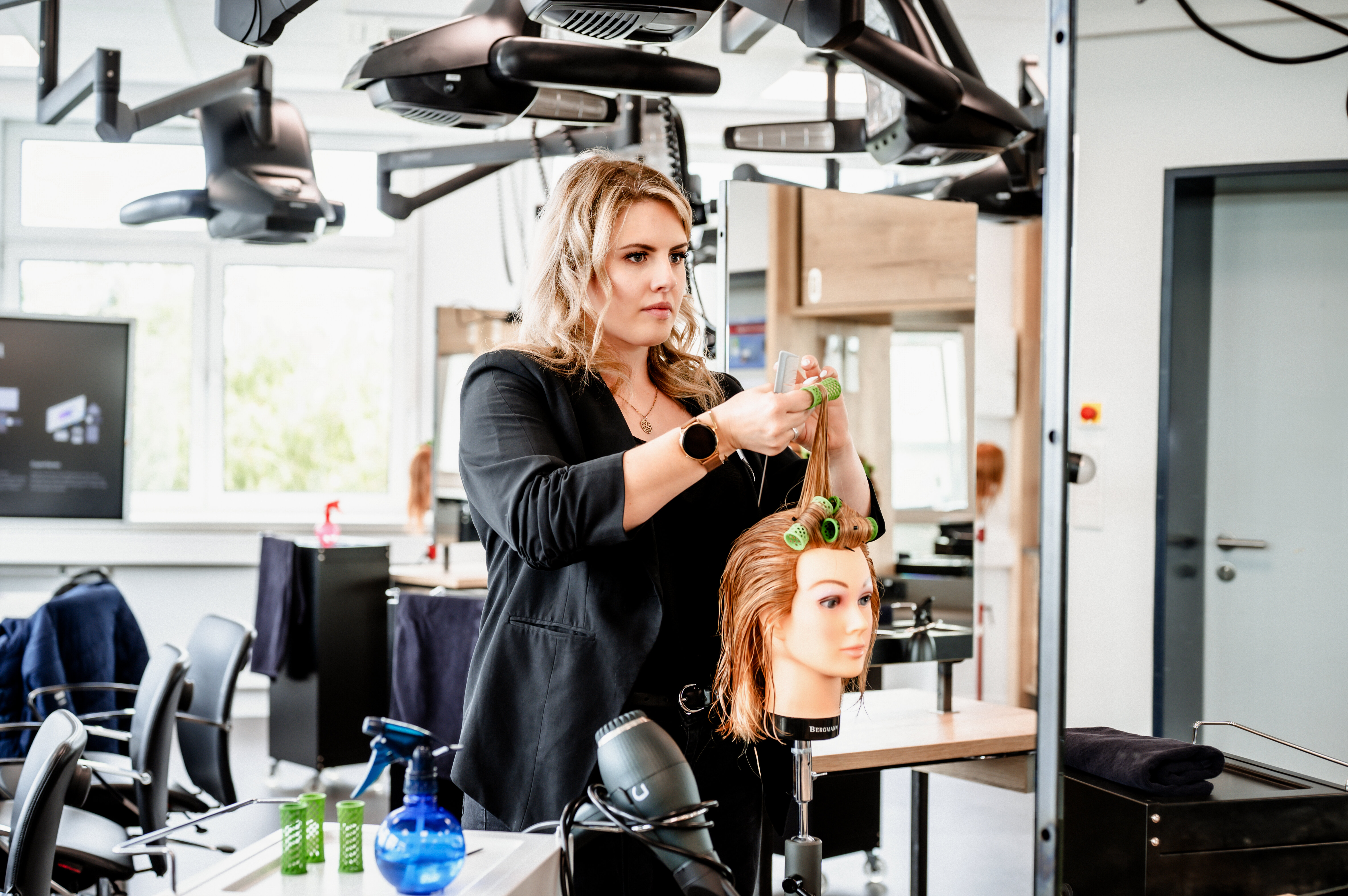 Eine Friseurin arbeitet konzentriert an einem Modellkopf mit Lockenwicklern in einem modernen Salon. Die Umgebung wirkt professionell und gut ausgestattet.