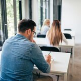 Ein Mann sitzt an einem Tisch in einem hellen Raum, scheinbar vertieft in seine Notizen, während andere Personen im Hintergrund sitzen.