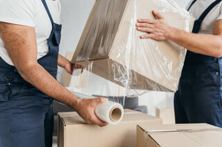 Zwei Männer in Blaumännern verpacken Pappkisten in Folie. 