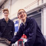 Das Bild zeigt zwei junge Menschen in Schutzanzügen und Schutzbrillen, die in einer Werkstatt an einer Maschine arbeiten. Sie lächeln freundlich.