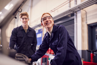 Das Bild zeigt zwei junge Menschen in Schutzanzügen und Schutzbrillen, die in einer Werkstatt an einer Maschine arbeiten. Sie lächeln freundlich.