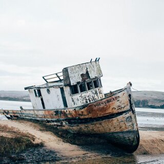 Ein altes, verfallenes Boot liegt auf dem Sand, teils im Wasser. Die umliegende Landschaft ist ruhig und leicht neblig.