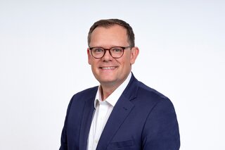 Adrian Burghardt mit Brille trägt einen blauen Blazer und ein weißes Hemd. Er lächelt freundlich vor einem neutralen Hintergrund.
