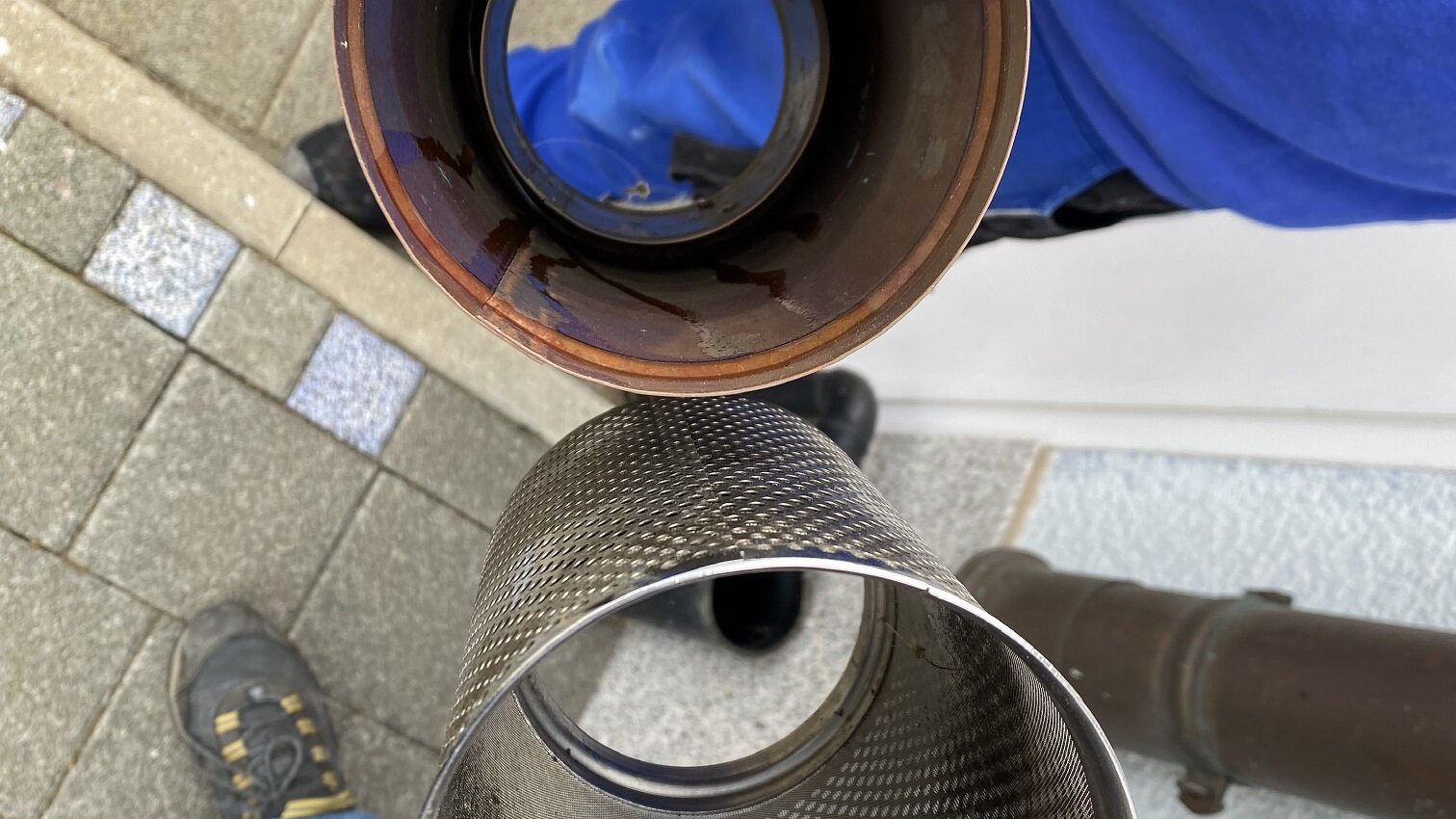 Das Bild zeigt zwei Auspuffrohre, eines mit Rost und das andere glänzend. Im Hintergrund sind Pflastersteine und Schuhe sichtbar.