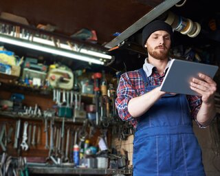 Ein Mann in einem blauen Overall und Beanie steht in einer Werkstatt, nutzt ein Tablet und umringt von Werkzeugen und Ausrüstung.