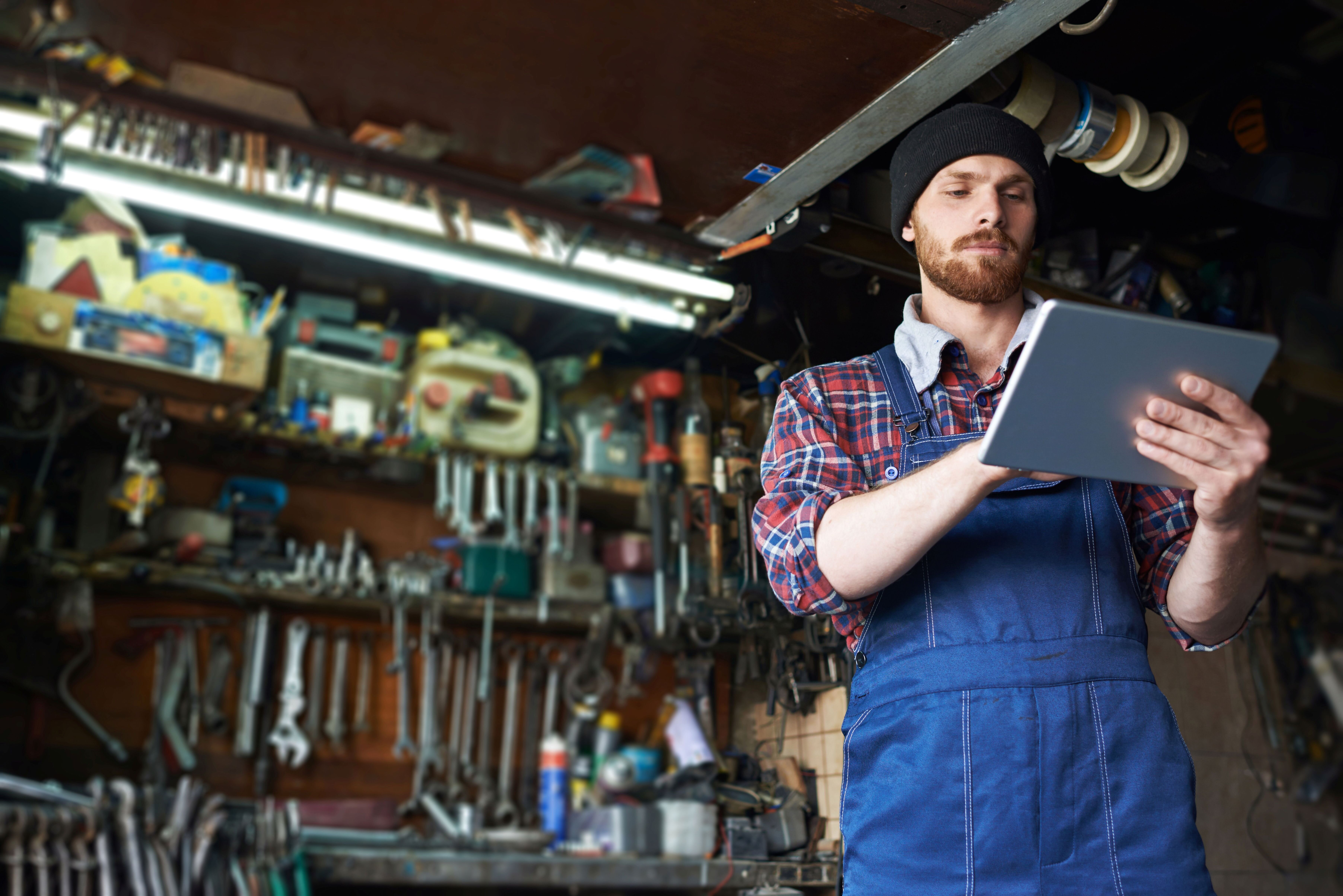 Ein Mann in einem blauen Overall und Beanie steht in einer Werkstatt, nutzt ein Tablet und umringt von Werkzeugen und Ausrüstung.