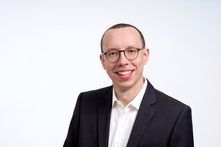 Ein lächelnder Mann mit Brille trägt einen schwarzen Anzug und ein weißes Hemd. Der Hintergrund ist hell und neutral.