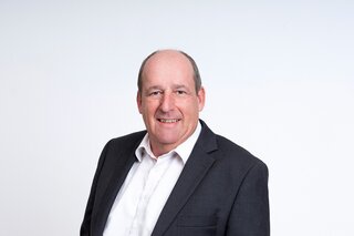 Ein lächelnder Mann mit Glatze trägt einen schwarzen Anzug und ein weißes Hemd. Der Hintergrund ist hell und neutral.