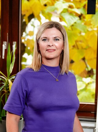 Eine Frau mit blonden Haaren trägt ein lila Oberteil. Im Hintergrund leuchten gelbe Blätter durch ein Fenster.