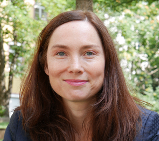 Das Bild zeigt Jeanette Pittmann, eine Frau mit langen, braunen Haaren und einem freundlichen Lächeln. Sie steht vor einer grünen, natürlichen Kulisse.