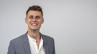Ein junger Mann mit kurzem, gelocktem Haar trägt einen grauen Blazer über einem weißen Hemd. Er lächelt freundlich vor einem neutralen Hintergrund.