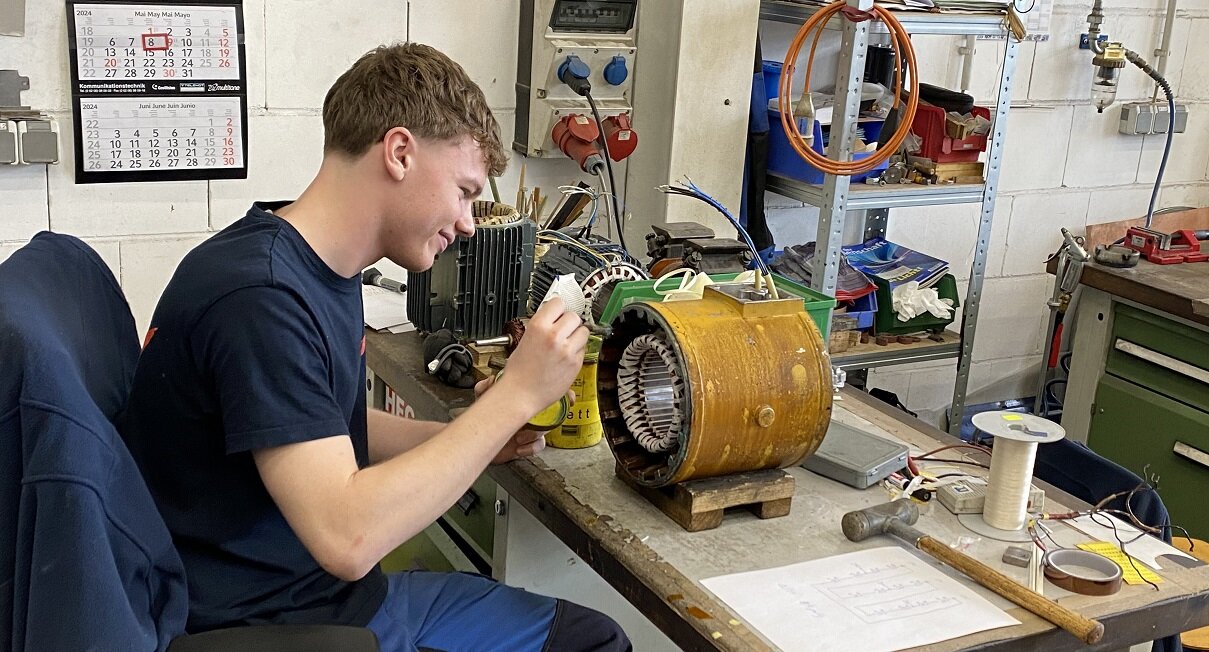Ein junger Mann arbeitet an einem Motor. Er ist konzentriert und verwendet Werkzeuge auf einem Tisch in einer Werkstatt. Im Hintergrund sind weitere Geräte.