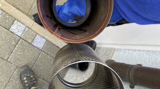 Das Bild zeigt zwei Auspuffrohre, eines mit Rost und das andere glänzend. Im Hintergrund sind Pflastersteine und Schuhe sichtbar.