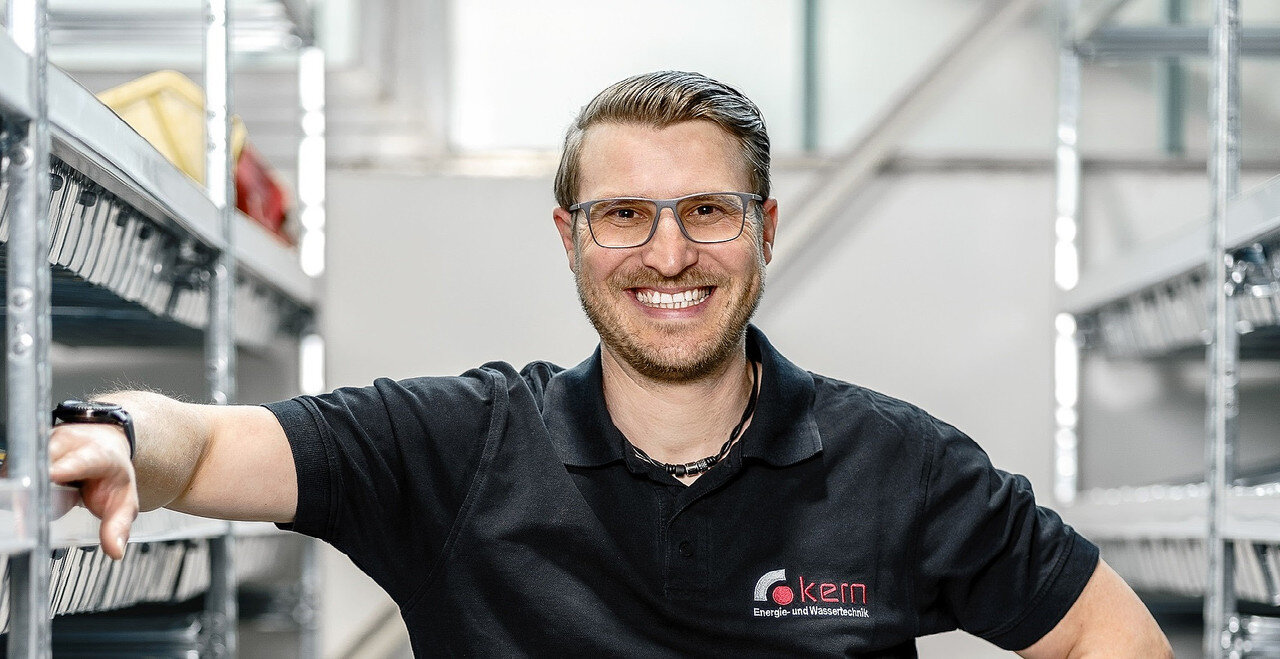 Ein lächelnder Mann in schwarzem Poloshirt steht entspannt zwischen Regalen, umgeben von Werkzeug und Technik.