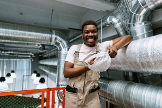 Ein junger Mann in Arbeitskleidung lächelt und hält eine Maske in der Hand. Im Hintergrund sind moderne Lüftungsrohre und Beleuchtung zu sehen.