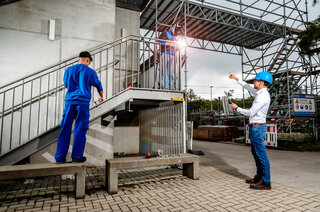 Das Bild zeigt zwei Bauarbeiter an einer Treppe auf einer Baustelle. Ein Arbeiter misst, während ein anderer Anweisungen gibt. Sicherheitsausrüstung ist vorhanden.