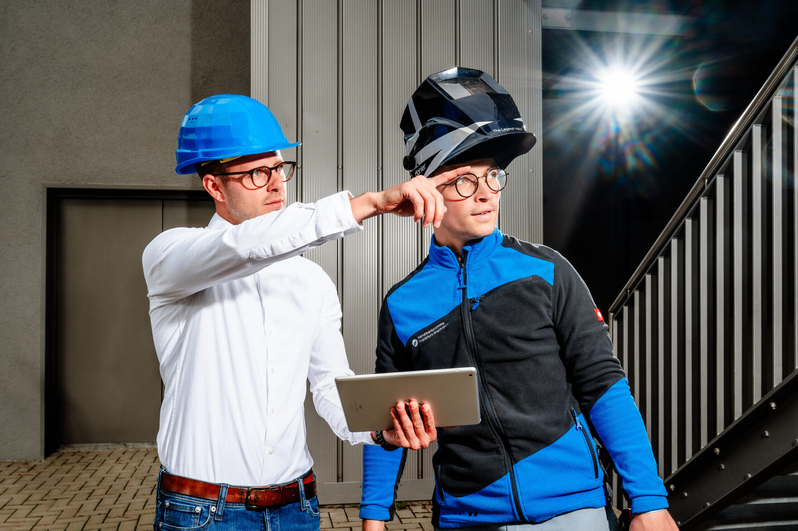 Zwei Männer in Sicherheitshelmen stehen im industriellen Umfeld. Einer zeigt auf etwas, während der andere ein Tablet hält.