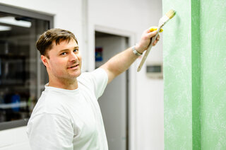 Ein Mann in einem weißen T-Shirt streicht eine grüne Wand. Er lächelt und hält einen Pinsel in der Hand.