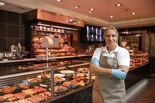 Die Szene zeigt eine freundliche Verkäuferin in einem gut sortierten Fleischereifachgeschäft. Die Theke ist reich gefüllt mit verschiedenen Fleischwaren und Delikatessen.