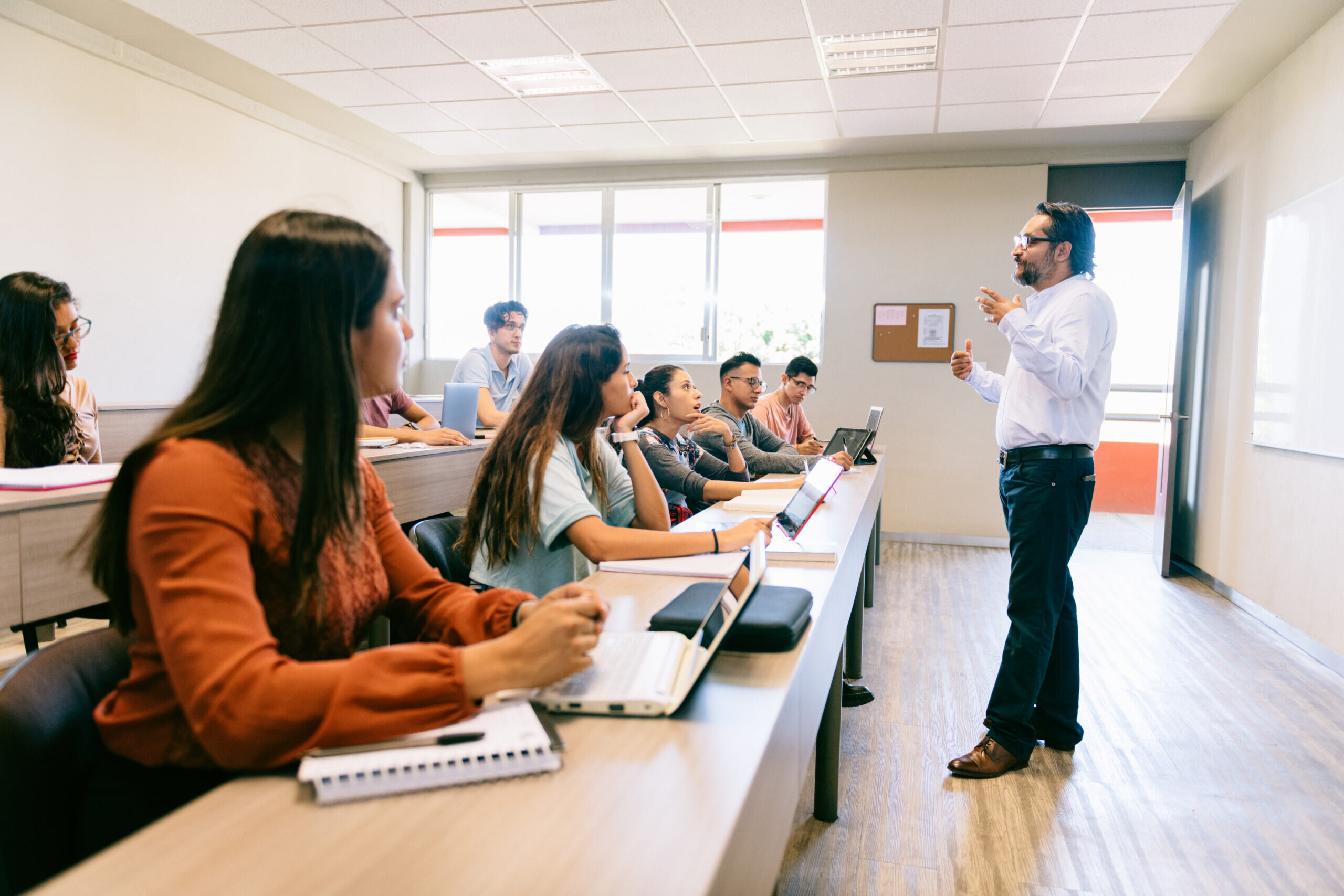 Ein Lehrer steht vor einer Gruppe von Studierenden in einem Klassenraum. Die Schüler arbeiten an Laptops und sind aufmerksam.