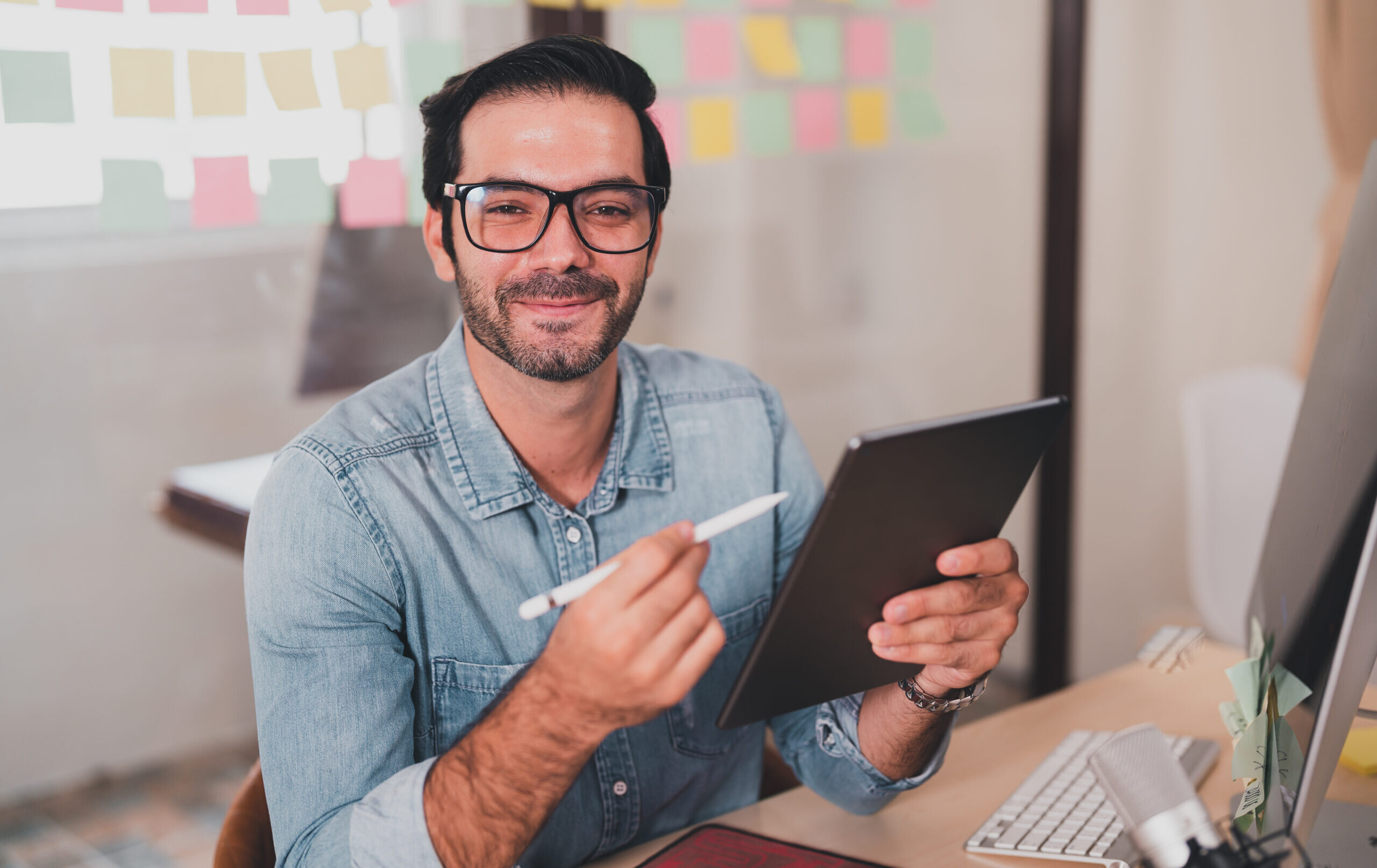 Ein lächelnder Mann mit Brille hält ein Tablet in der Hand. Er trägt ein Jeanshemd und sitzt an einem Schreibtisch mit bunten Notizen im Hintergrund.