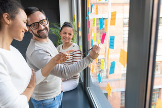 Drei Personen stehen an einem Fenster und arbeiten gemeinsam an bunten Haftnotizen. Sie lachen und scheinen sich aktiv auszutauschen.
