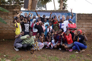 Eine Gruppe junger Menschen steht fröhlich vor einer Wand, zeigt Peace-Zeichen und lächelt. Die Stimmung ist herzlich und einladend.