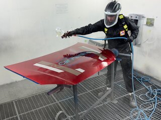 Ein Arbeiter in Schutzkleidung sprüht rotlackierte Autoteile in einer Lackiererei. Die Umgebung ist sauber und gut beleuchtet.