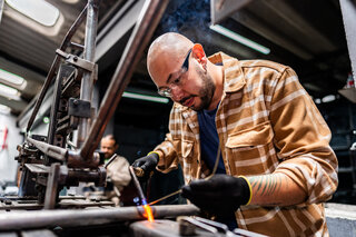 Ein Handwerker arbeitet konzentriert an einem Metallprojekt. Er trägt Schutzbrille und Handschuhe, während eine Flamme aufleuchtet.