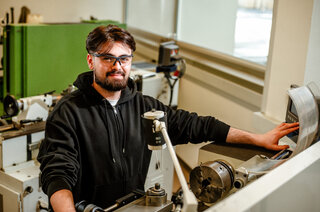 Ein junger Mann mit Brille lächelt am Arbeitsplatz in einer Maschinenwerkstatt. Er trägt einen schwarzen Hoodie und steht neben einer Drehmaschine.